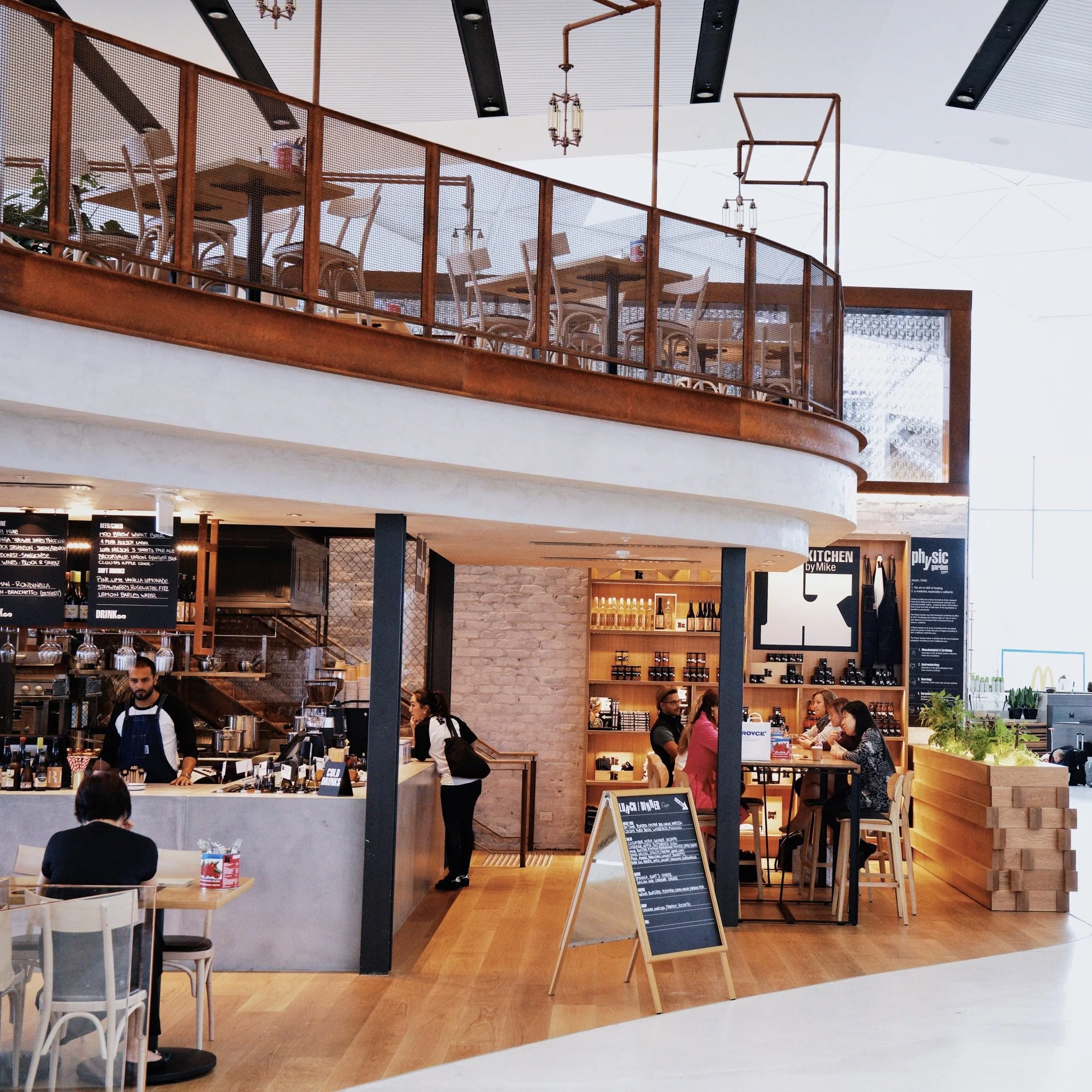 Inside a modern café or coffee shop with a second-floor seating area, a barista preparing drinks behind the counter, and customers seated at tables.
