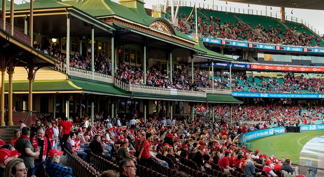 Crowd of baseball spectators seated in a stadium stands, many wearing team colors and apparel, with some standing and talking, during a game.