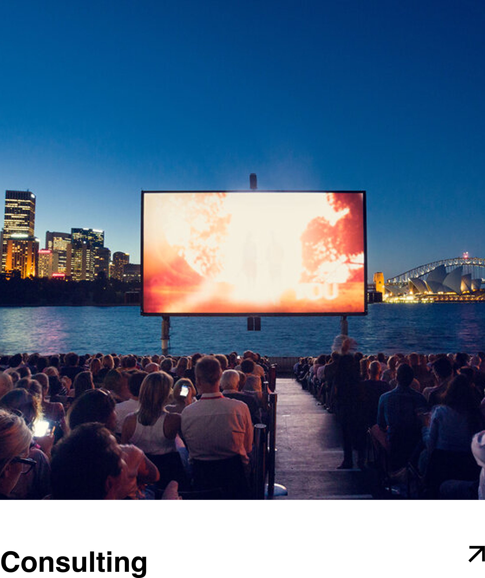 People watching an outdoor movie on a large screen by the water during evening, with city skyline and Sydney Opera House in background.
