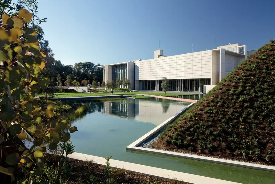 Modern building with extensive glass windows, surrounded by greenery, with a reflective pond and landscaped garden in front.