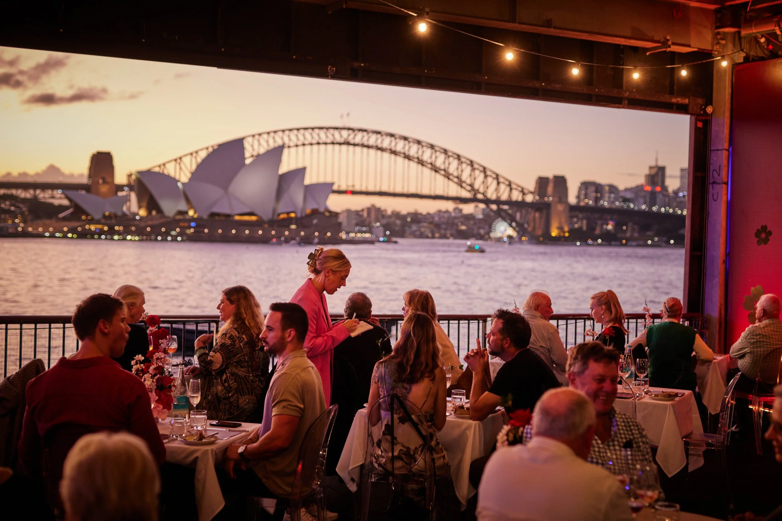 People dining at a restaurant with a view of Sydney Opera House and Harbour Bridge during sunset.