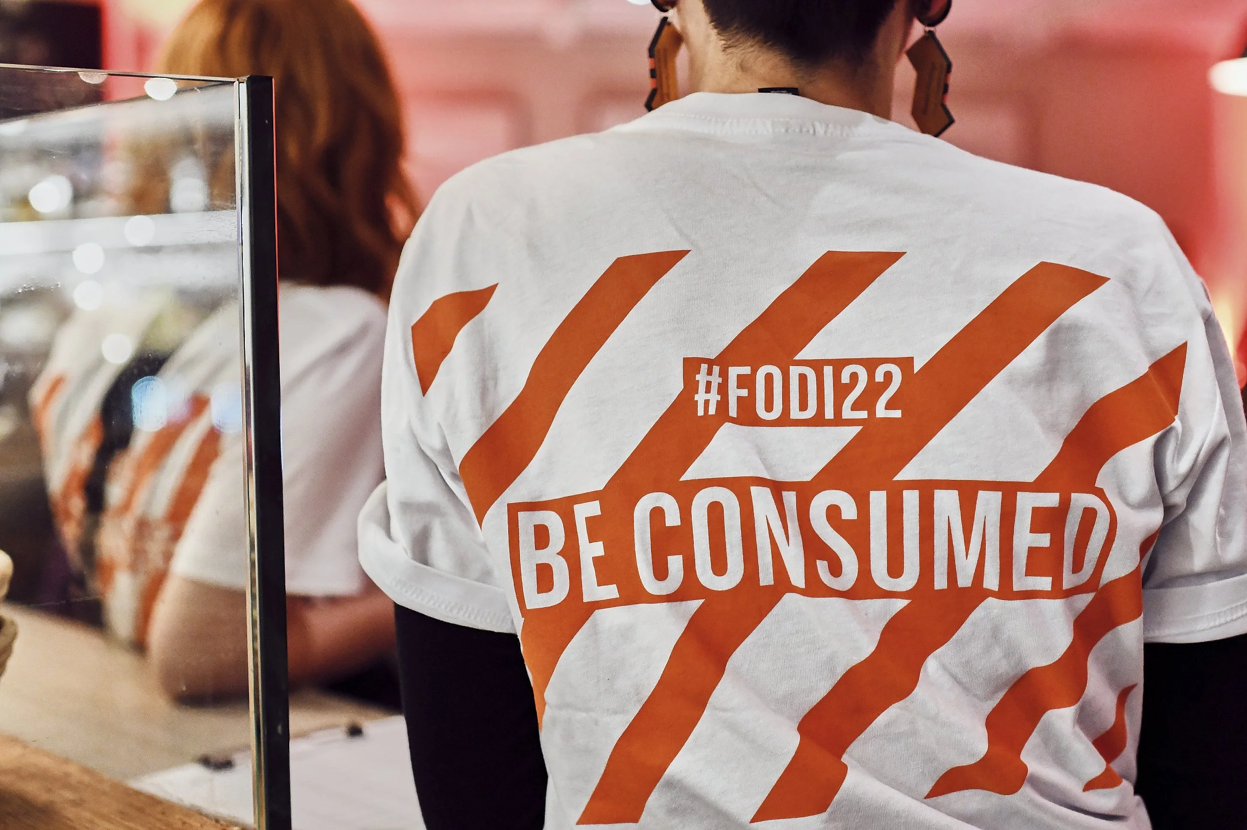 Back of a person wearing a white t-shirt with orange diagonal stripes and bold text that reads '#FODI22 BE CONSUMED' in a busy indoor setting with other people and a transparent divider.