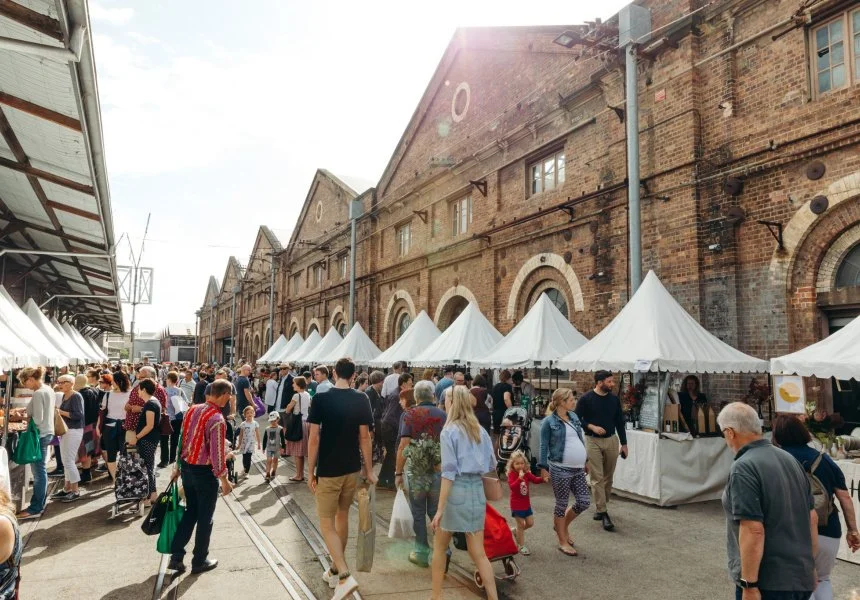 Outdoor market with white tents and a crowd of people shopping, set against historic brick buildings on a sunny day.