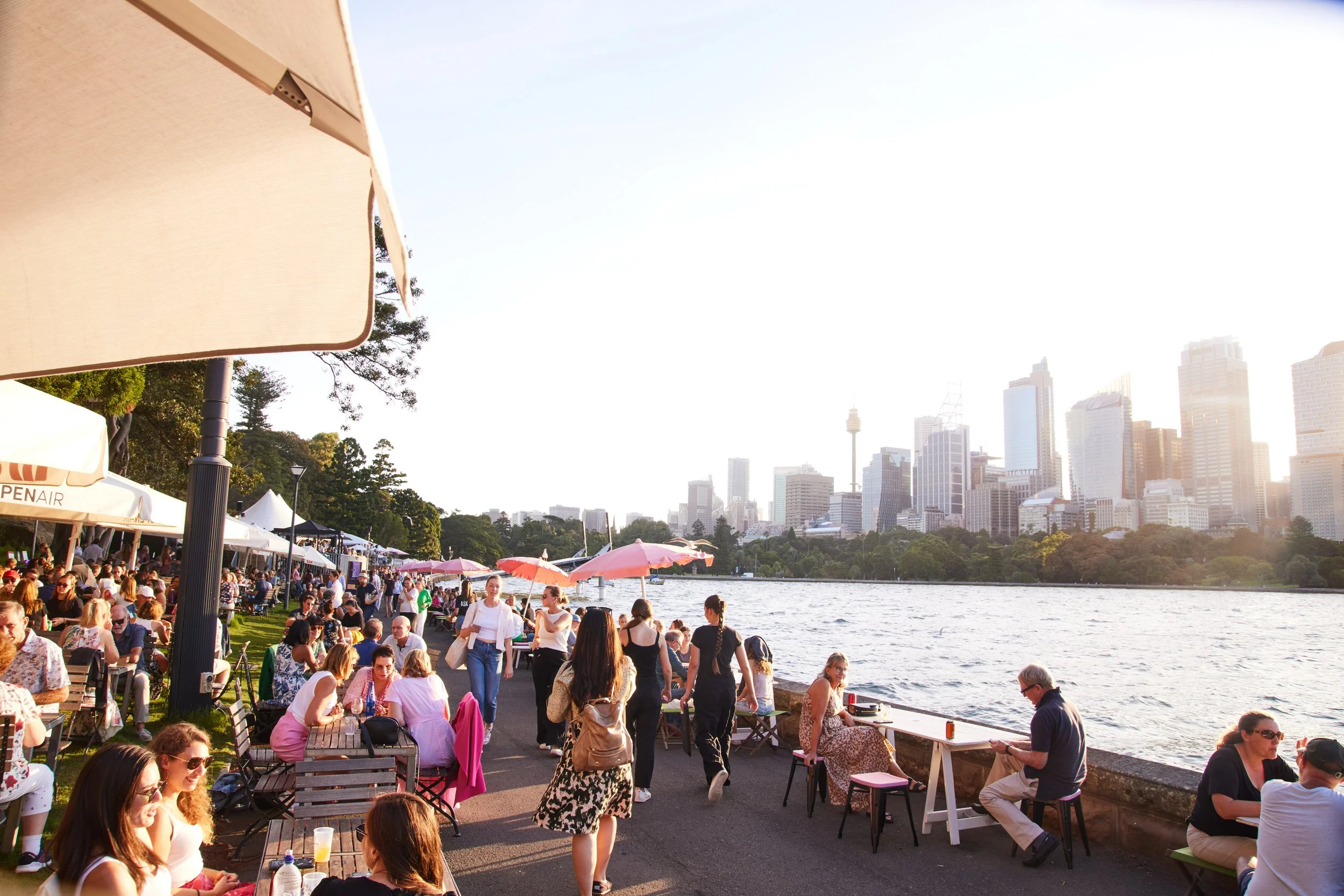 People dining and socializing along a riverside promenade with city skyscrapers in the background during daytime.