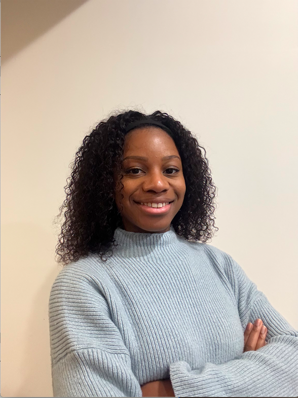 A woman with dark curly hair, wearing a gray sweater, smiling with arms crossed in front of a plain wall.