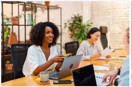 Two women sitting at a desk in a modern office, working on laptops and reading documents, with potted plants and shelves in the background.