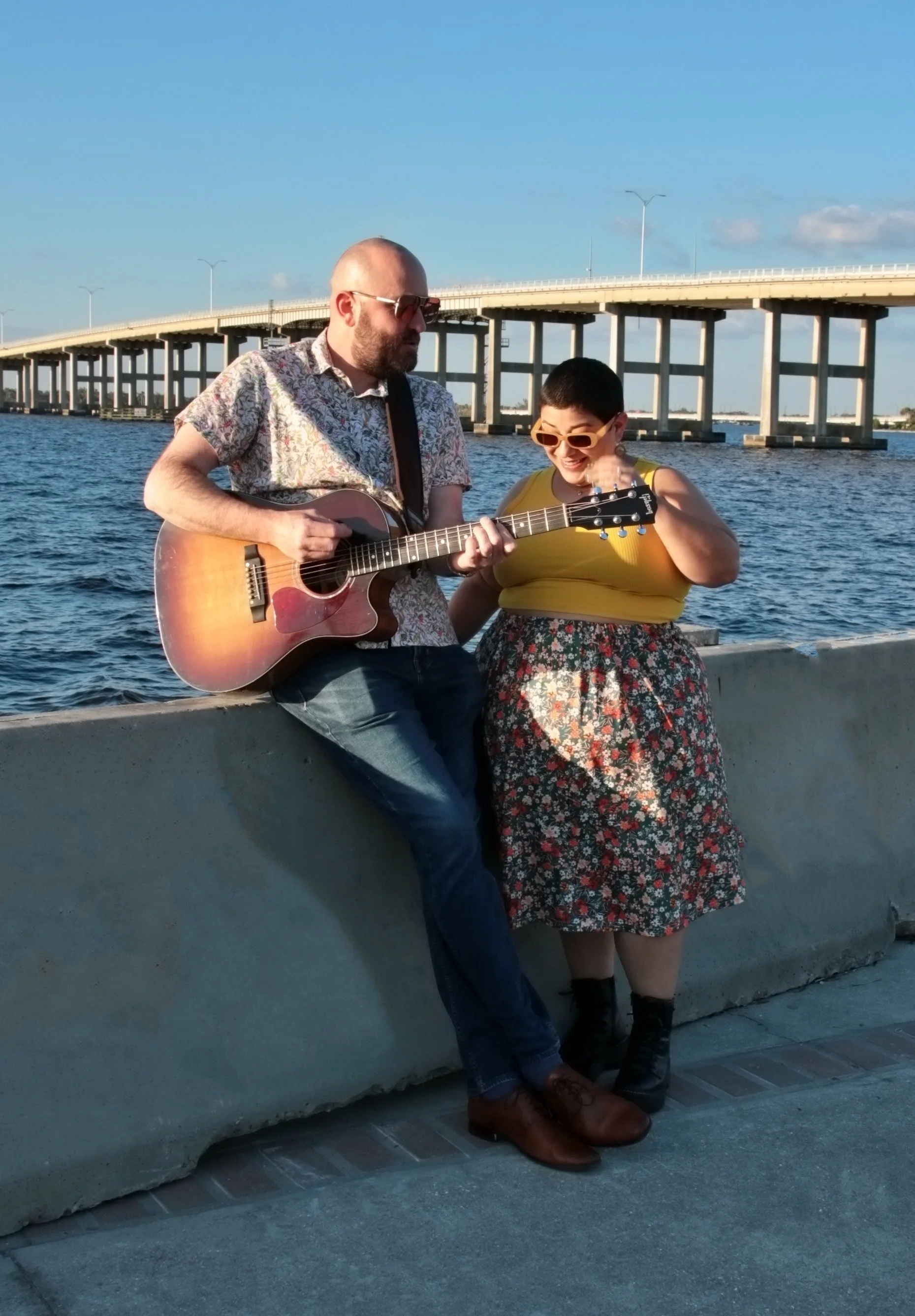 Two people, a man and a woman, sitting near a body of water with a bridge in the background. The man is playing an acoustic guitar while the woman is smiling and looking at him. The scene appears to be in the late afternoon with clear skies.