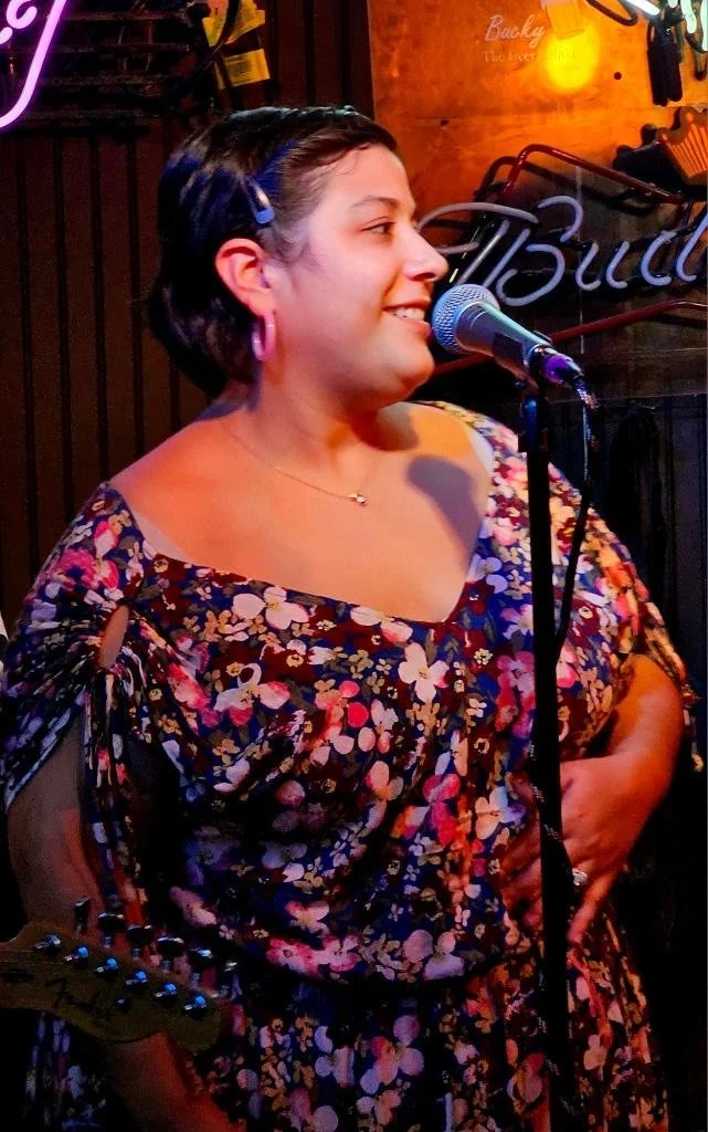 A woman singing into a microphone at a bar or club, wearing a floral dress, with headphones on and short dark hair.