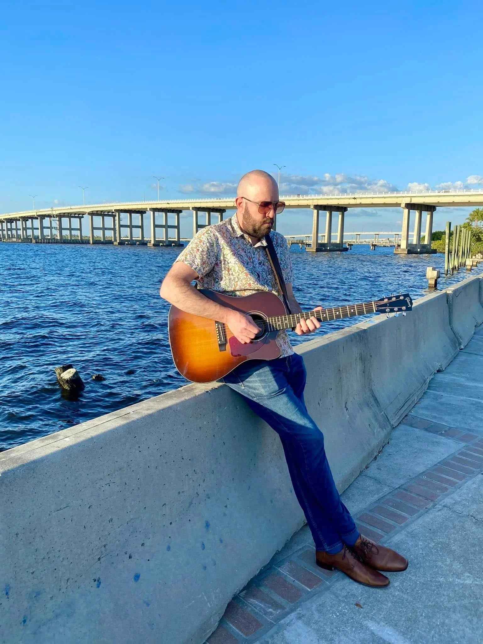 A man wearing sunglasses, a patterned shirt, blue jeans, and brown shoes is sitting on a concrete barrier by the water, playing an acoustic guitar. A bridge is visible in the background under a blue sky with some clouds.