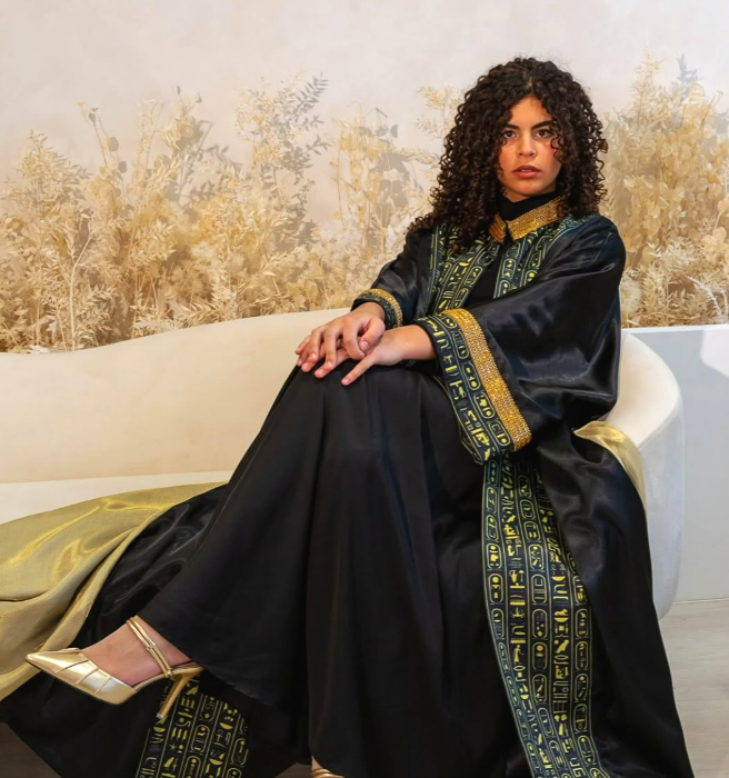 A woman with curly hair wearing a black and gold embroidered robe sitting on a white curved sofa, with a gold patterned fabric draped over her legs and gold heels. Behind her is a wall with a nature-themed mural or wallpaper.