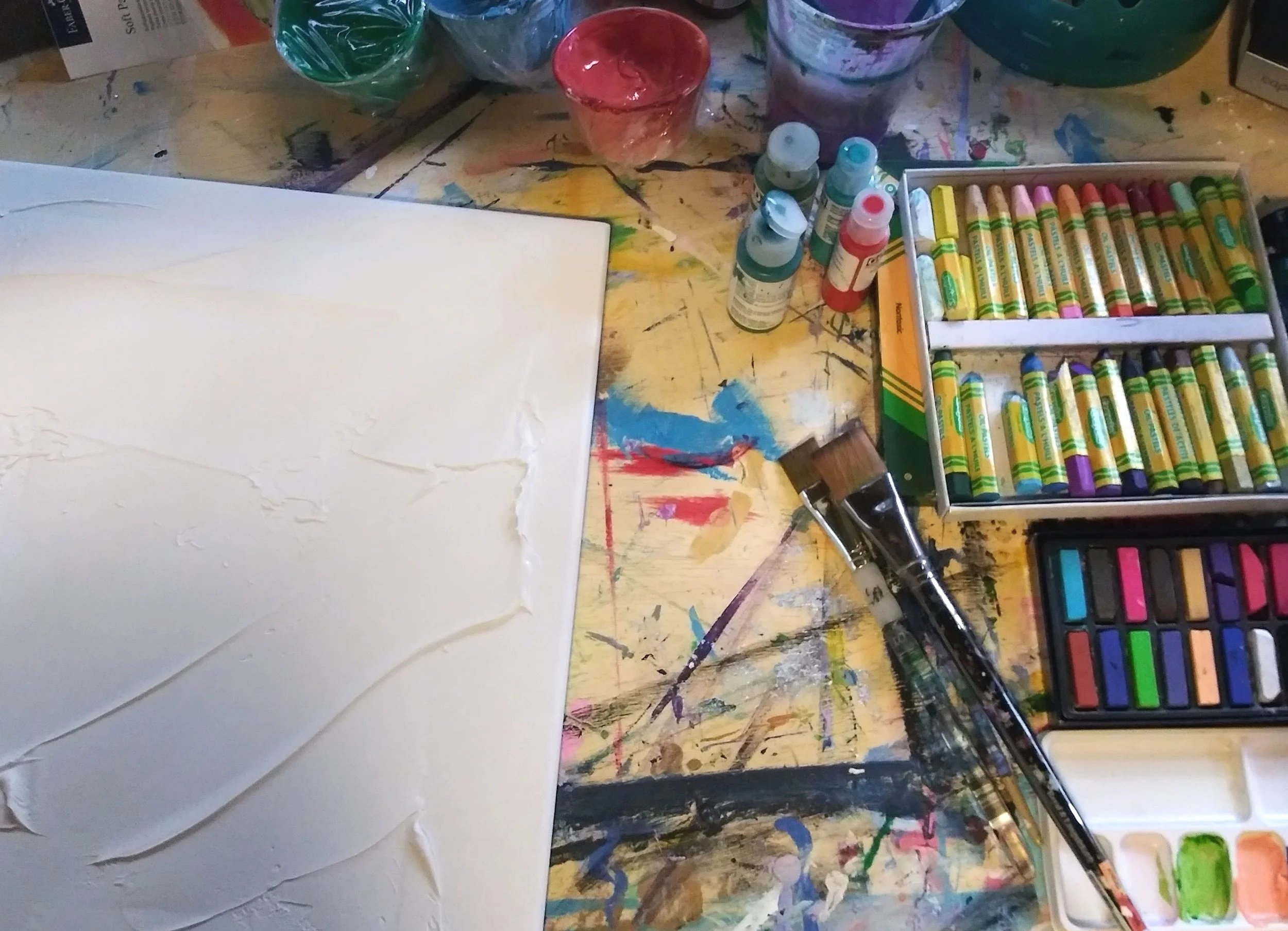 Art supplies on a cluttered artist's workspace: tubes of acrylic paint, paintbrushes, watercolor set, jars of paint, and an empty canvas with textured white paint, all arranged on a splattered, multicolored table.