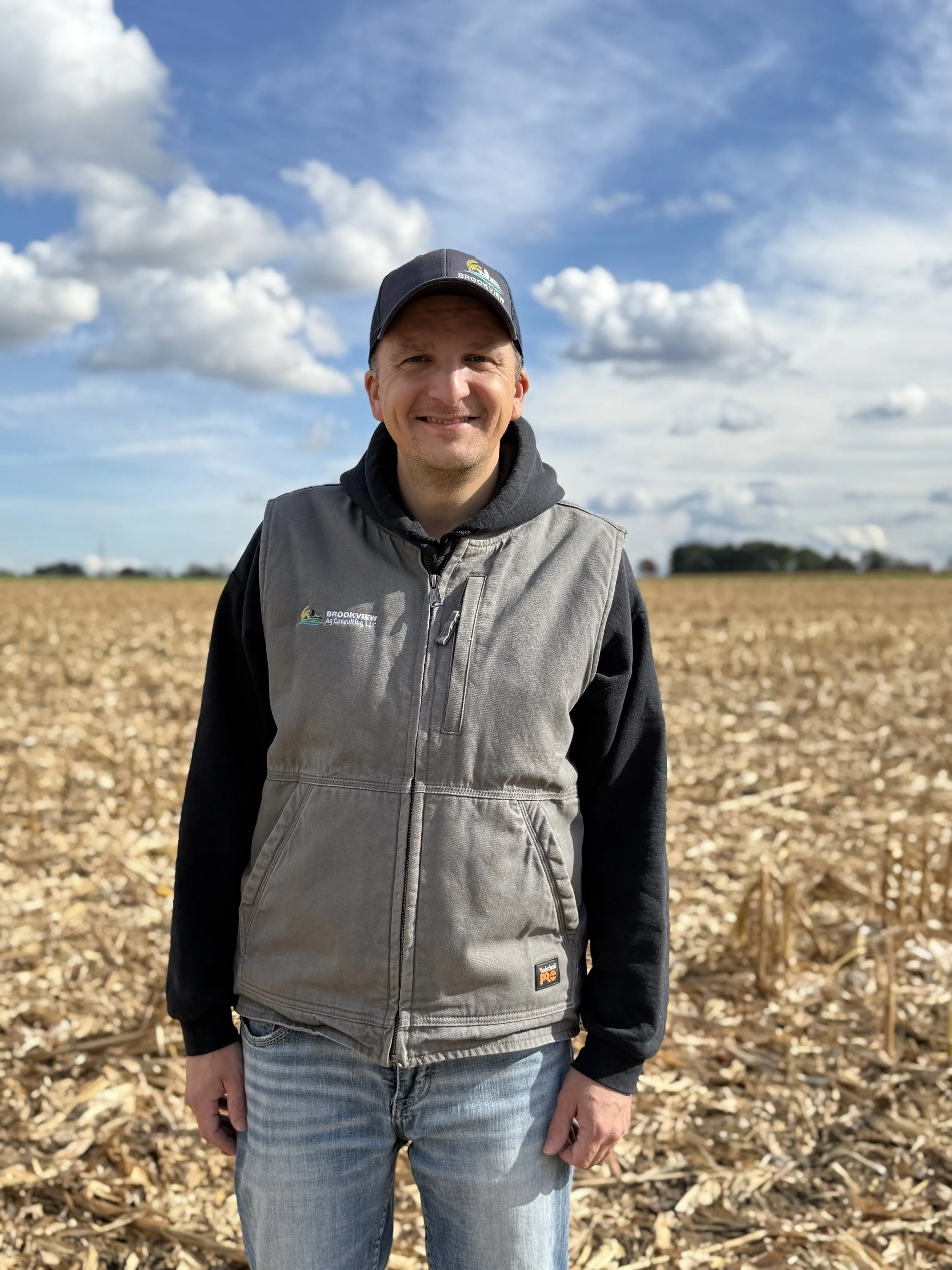 A man standing outdoors in a harvested cornfield with a partly cloudy sky behind him, wearing a Brookview agricultural vest and a cap.
