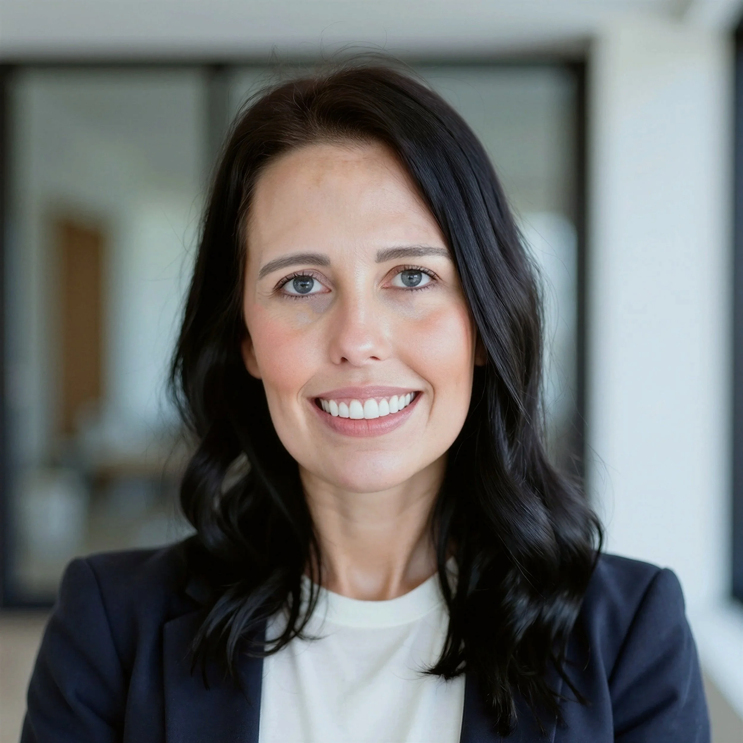 Close-up of a woman with blue eyes and black hair, smiling, wearing a dark blazer and a white shirt, standing indoors.