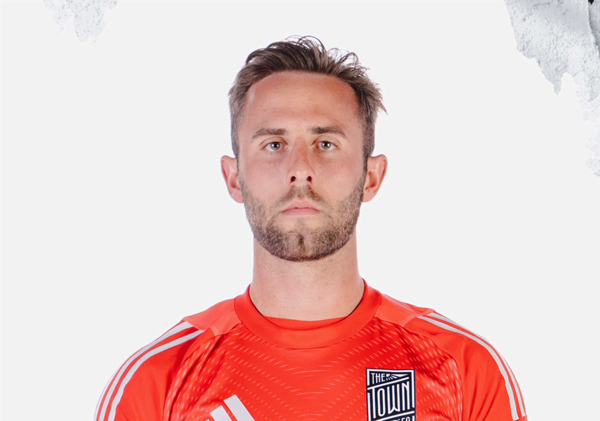 A man with short, styled hair and a beard is wearing a red sports jersey with white stripes and a badge that reads "The Town" on a plain white background.