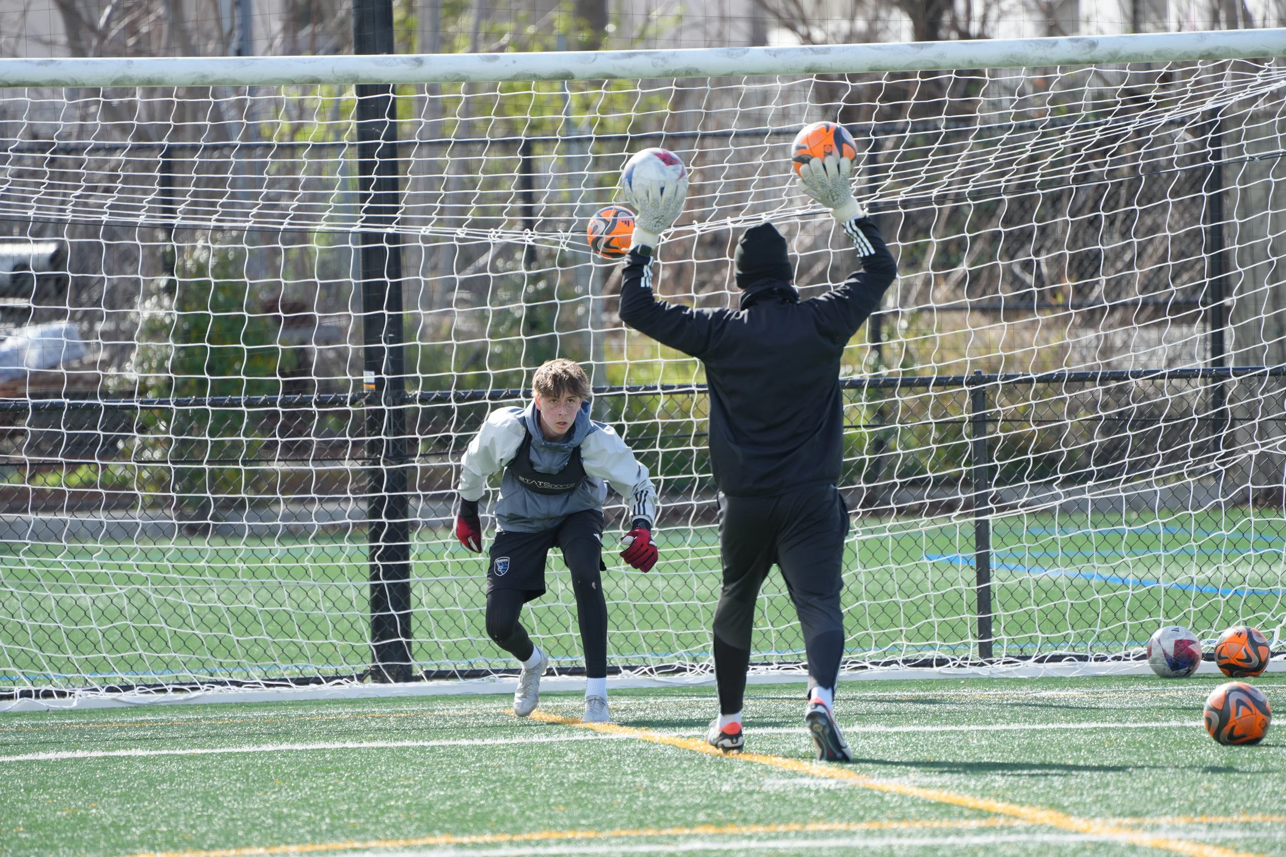 Two soccer players practice goalkeeping on a field with a net, one holding a ball in each hand and the other ready to catch a shot.