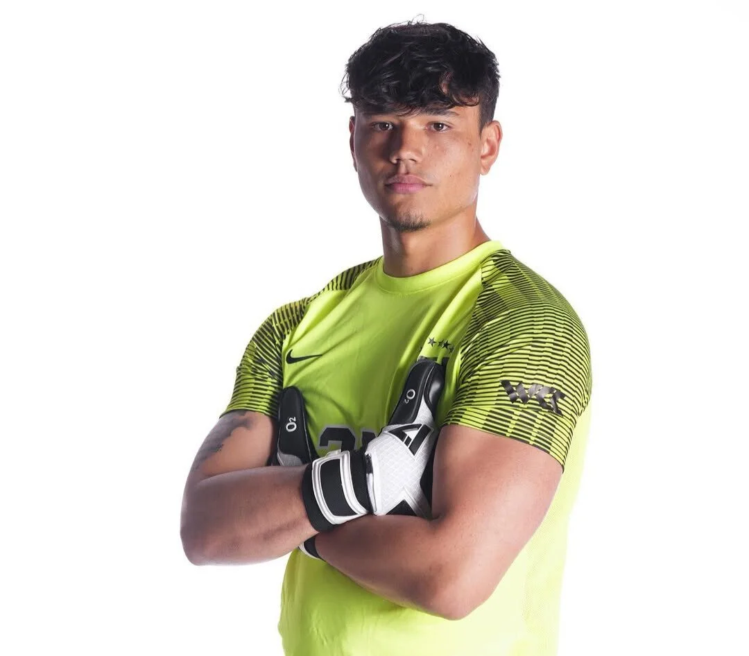 A male soccer goalkeeper wearing a neon yellow jersey and gloves, standing with arms crossed against a white background.