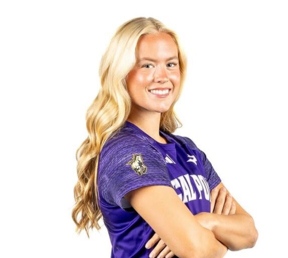 A young woman with long blonde hair wearing a purple sports jersey, smiling, with arms crossed.