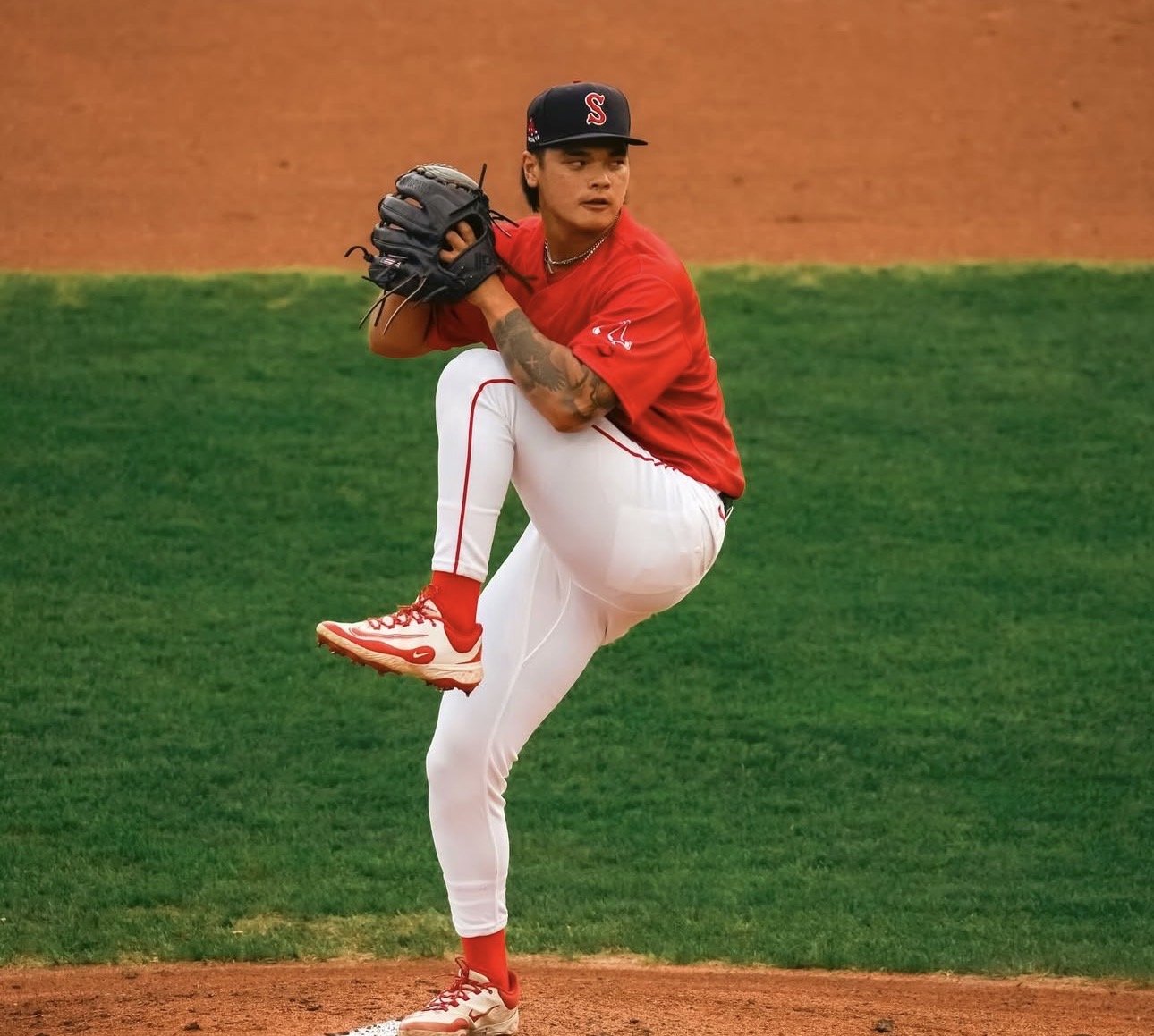 A baseball pitcher in a red jersey and white pants is in the middle of a pitch, with one leg raised and a glove on his hand on the pitcher's mound.