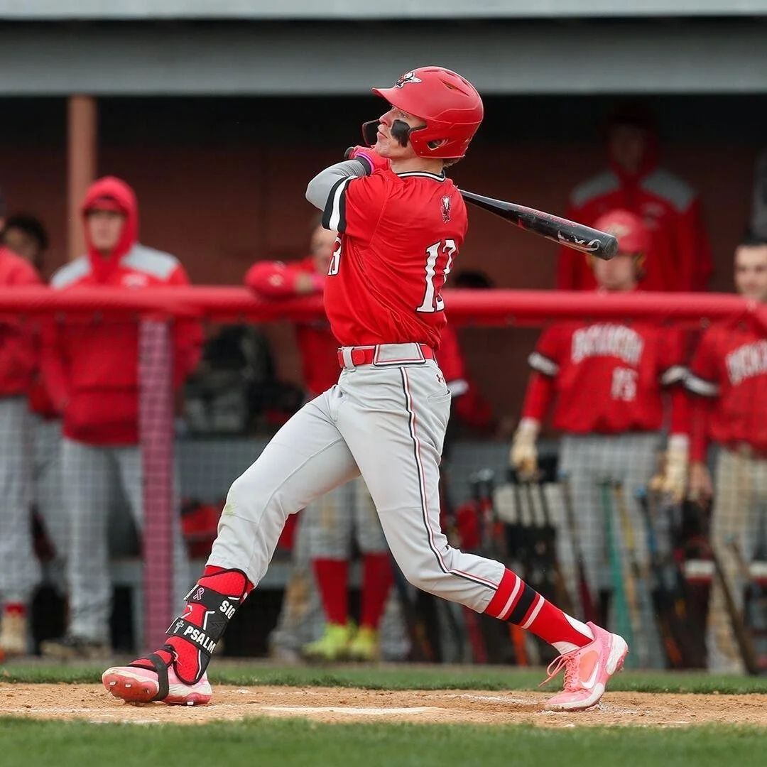 A baseball player in a red uniform and helmet is swinging a bat during a game, with teammates and coaches in the background.