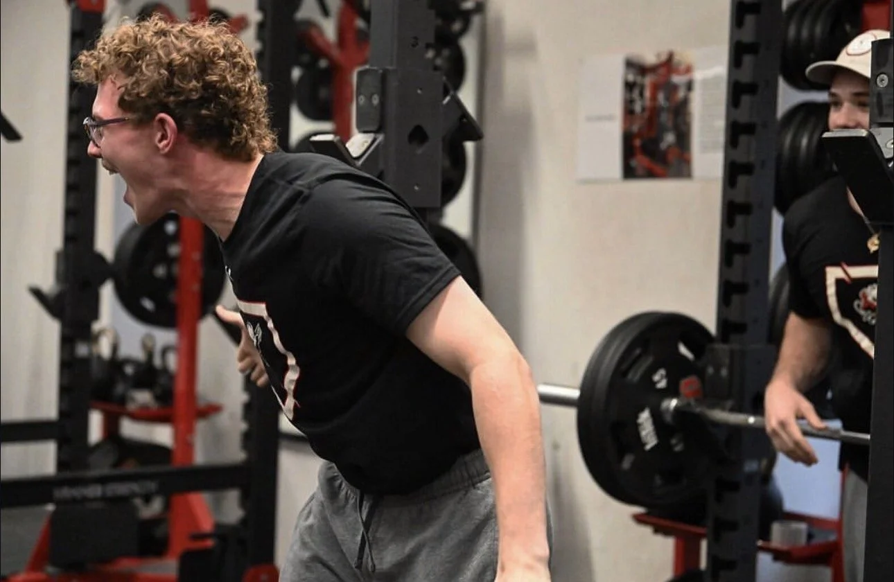 A young man with curly hair and glasses lifting a barbell during a workout in a gym, with another person standing nearby.