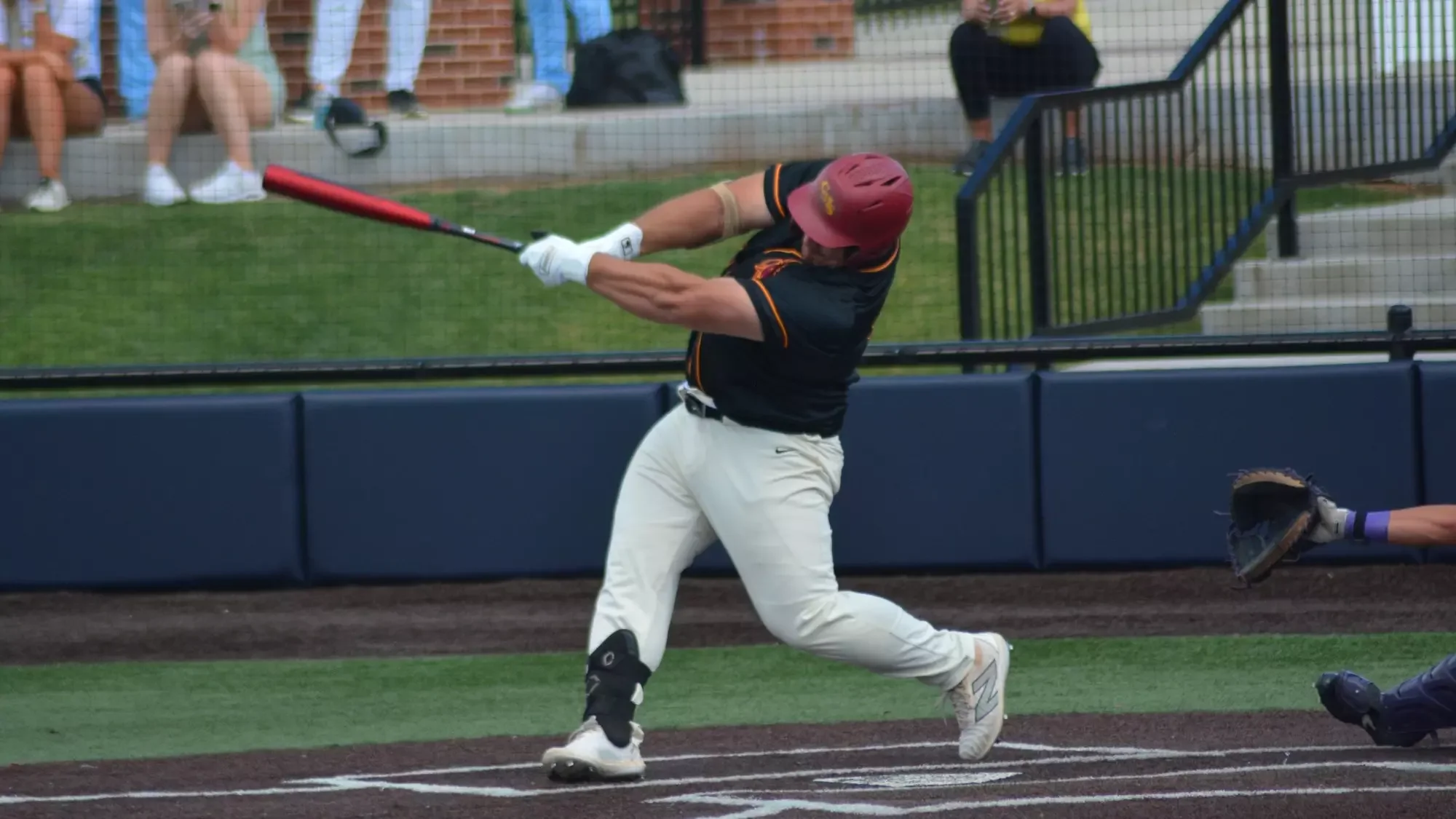 A baseball player wearing a maroon helmet, black jersey, and white pants is swinging a bat at a pitched ball on the field. The player is in a right-handed stance, with a catcher behind him and spectators watching from the stands in the background.