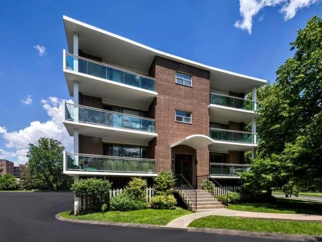 Modern four-story apartment building with brick facade, glass balconies, and a curved roof over the entrance, surrounded by greenery and trees.