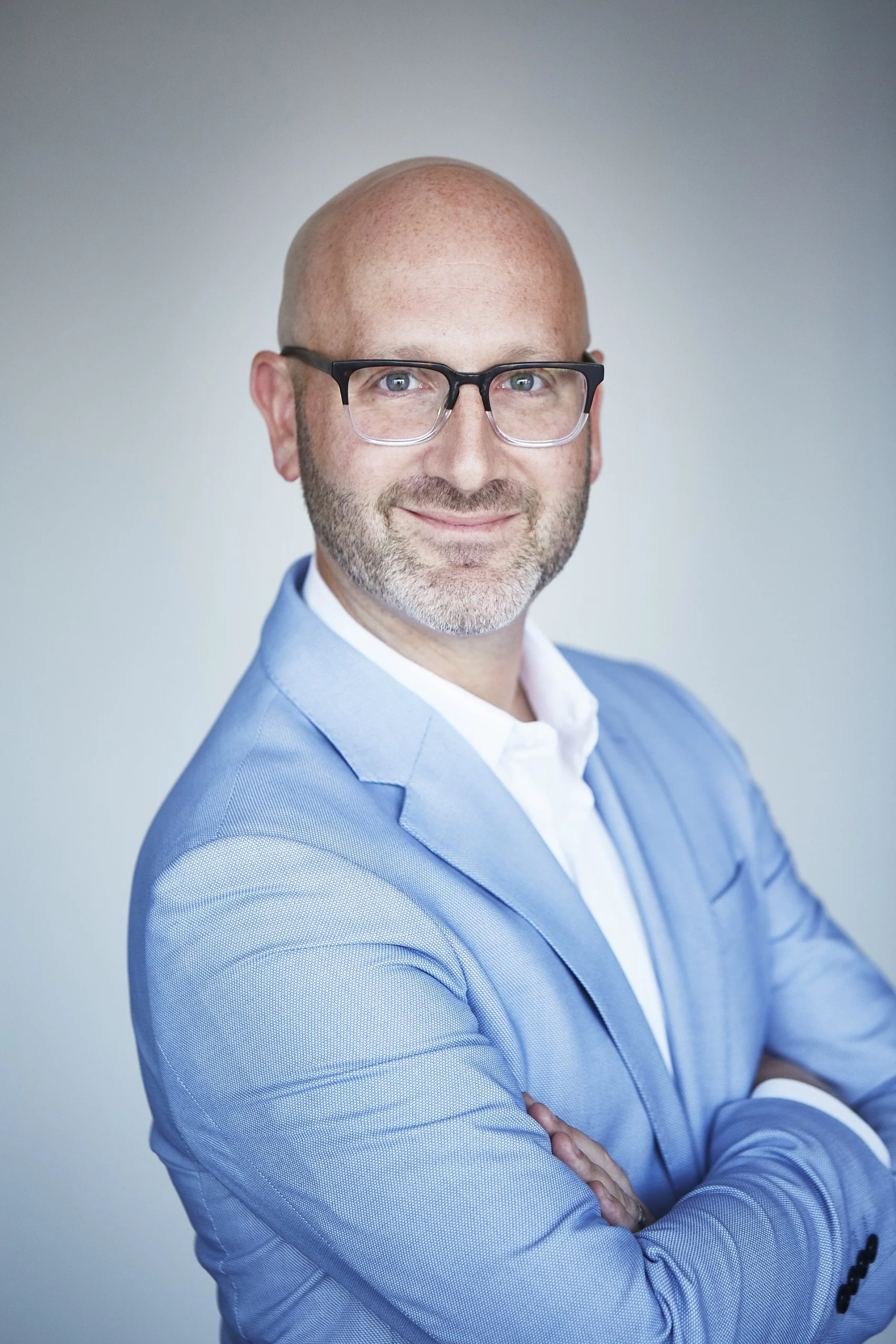 A professional man with glasses, a bald head, and a trimmed beard, smiling with arms crossed, wearing a light blue suit jacket and a white shirt against a neutral background.