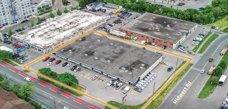 Aerial view of a parking lot with several commercial buildings, cars parked in and around, and streets labeled Midwest Rd and another unnamed road in a city area.