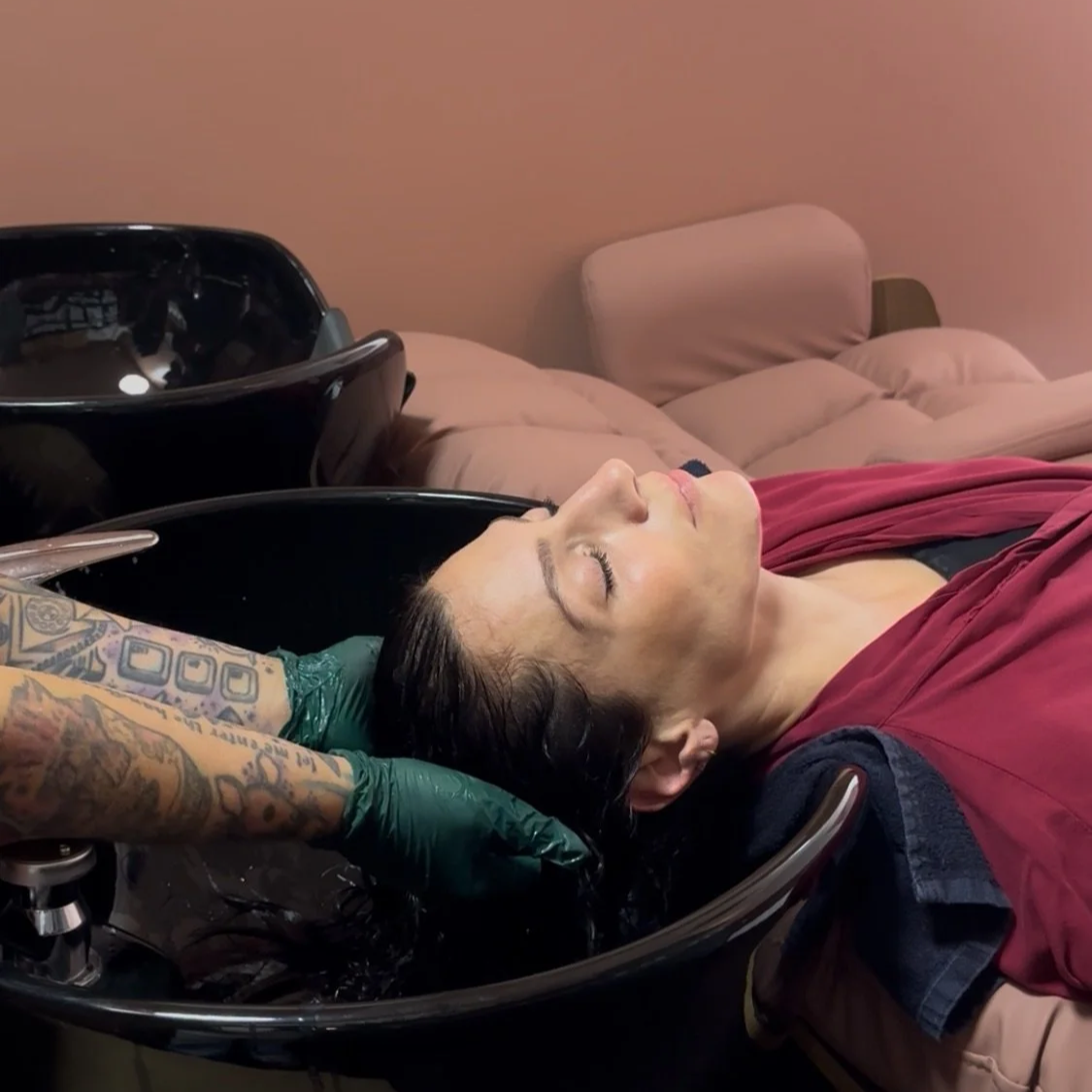 A woman with dark hair receives a hair wash at a salon, reclining with her eyes closed, wearing a maroon salon cape, as tattooed hands wash her hair in a black basin, with a beige bed and wall in the background.