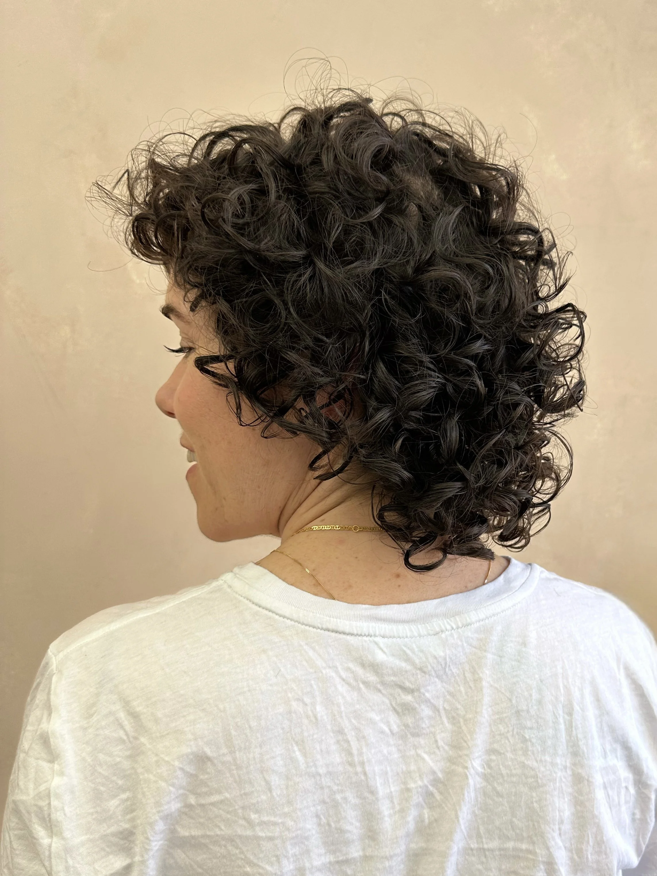 Profile view of a woman with short, dark curly hair, wearing a white top and a delicate gold necklace, standing against a neutral wall background.