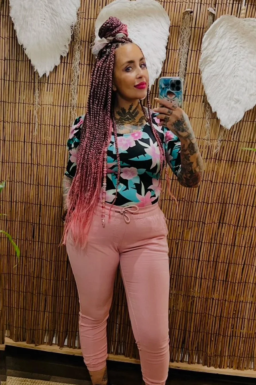 A woman with long pink braids taking a mirror selfie in front of a bamboo wall with large white heart-shaped decorations.