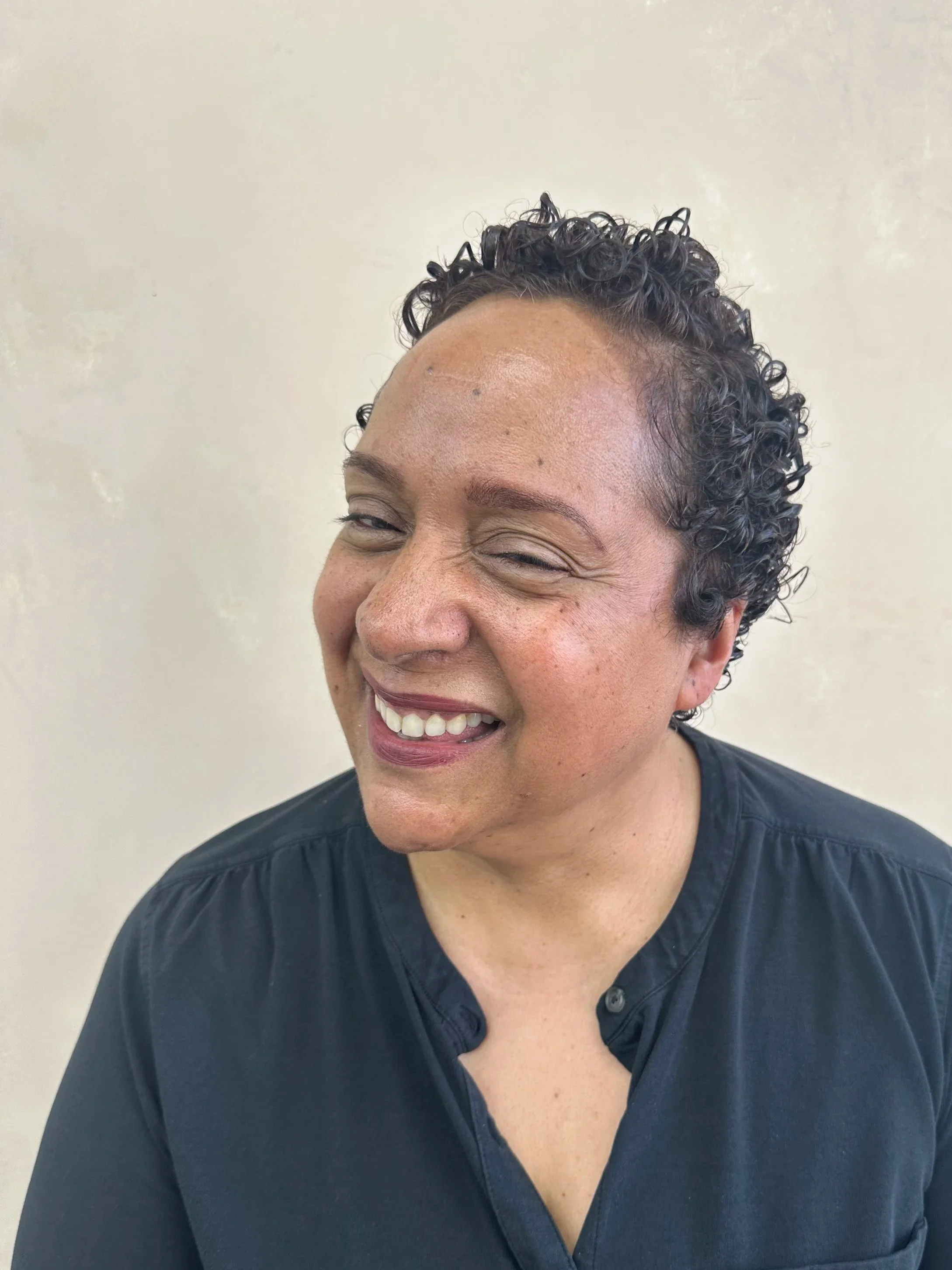 Close-up of a smiling woman with short, curly dark hair, wearing a black shirt, visible from her shoulders up against a plain light-colored background.
