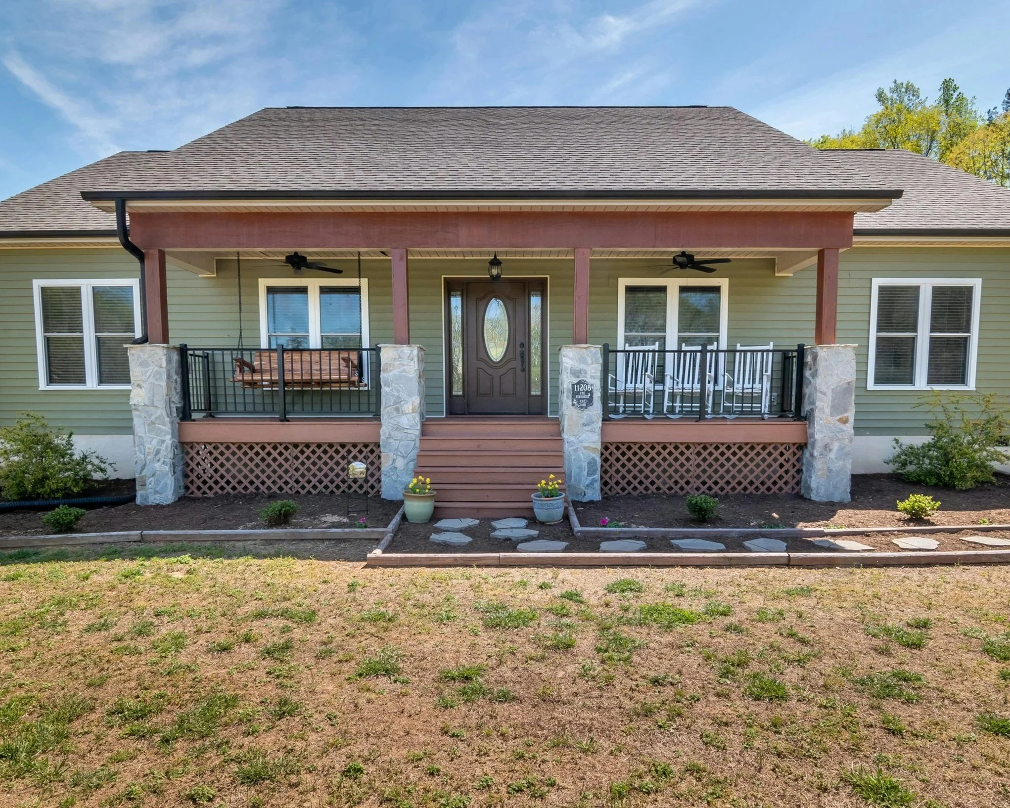 photo of the front of a home where the door is a dark wood, the house is painted green, the front porch is a light colored wood, the overhang a reddish brown, and the rock columns light grey. The colors draw the eye to different parts of the house.