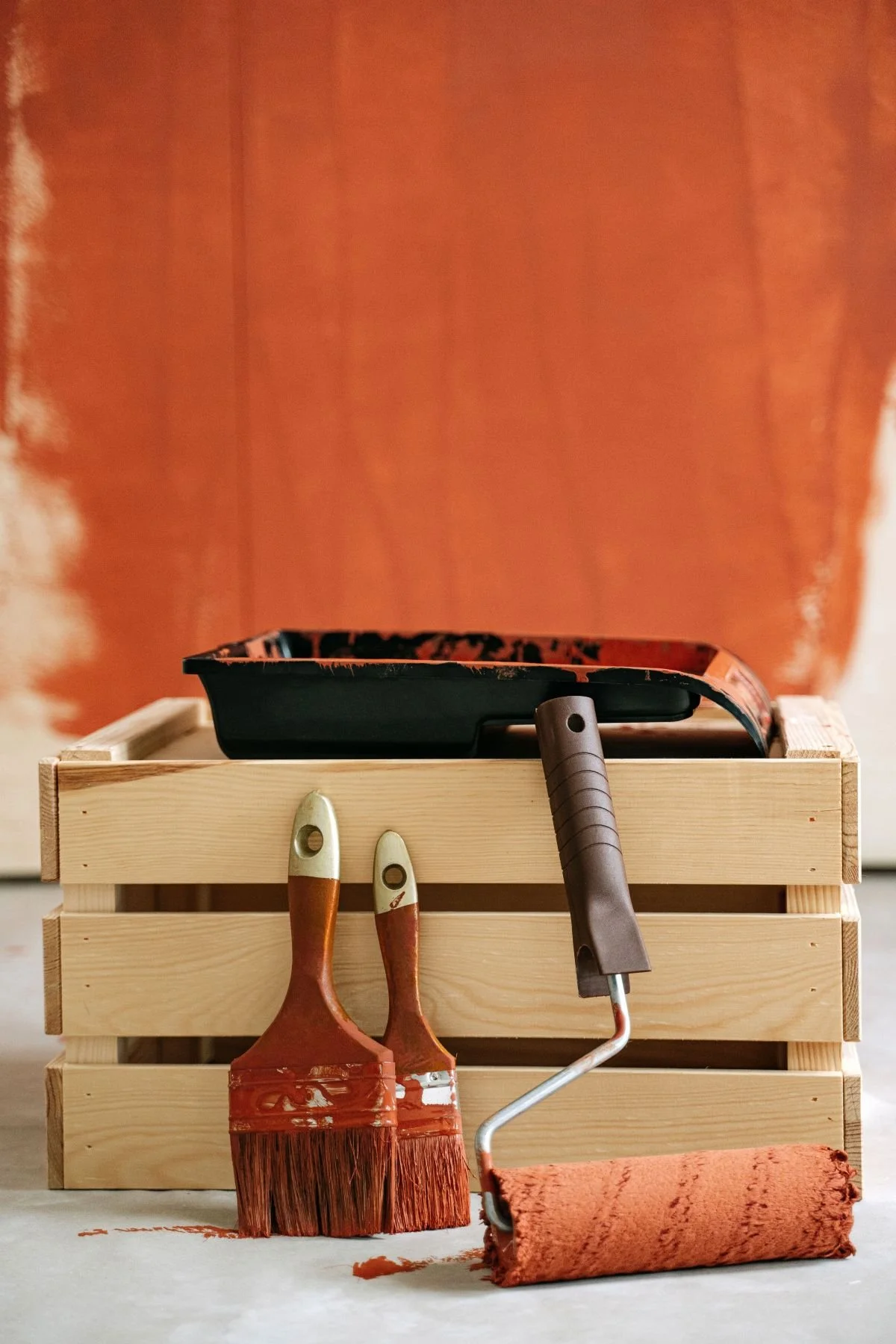 a wet paint tray sits atop a wooden crate in front of an orange painted wall. In front of the crate rests two paintbrushes and a paint roller all soaked with the same orange paint