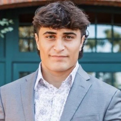 Young man with dark hair wearing a gray suit and white patterned shirt, standing outdoors in front of a building with large windows.