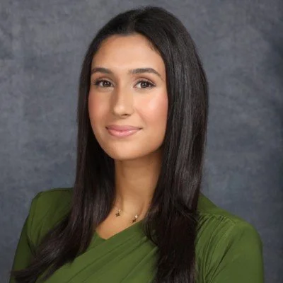 Portrait of a woman with long dark hair wearing a green top, standing against a gray background.