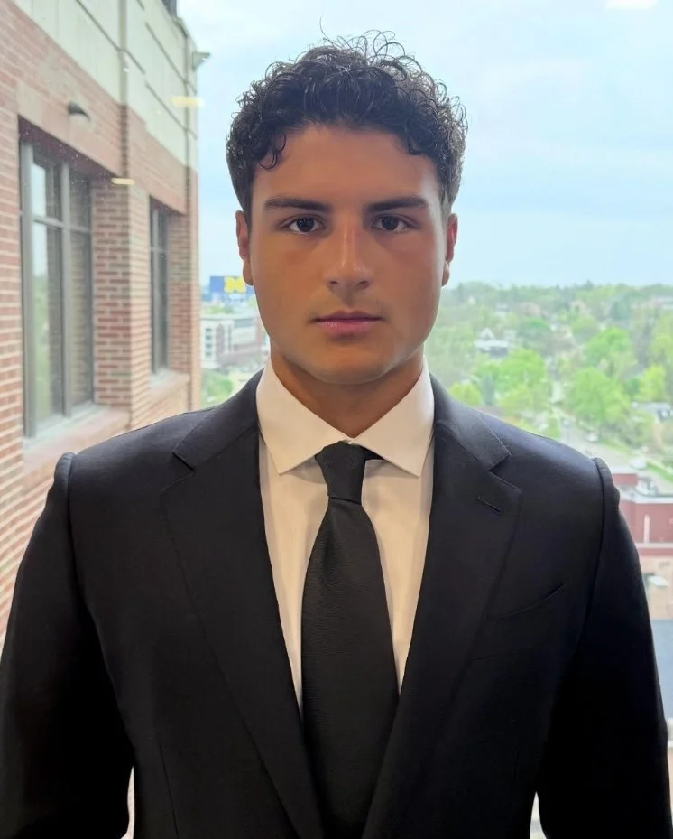 A young man in a black suit and tie standing indoors near a window with a cityscape in the background.