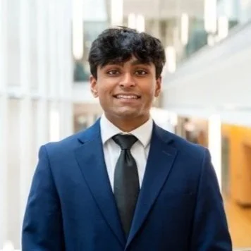 A young man in a navy blue suit and tie smiling in an indoor setting with modern architecture.