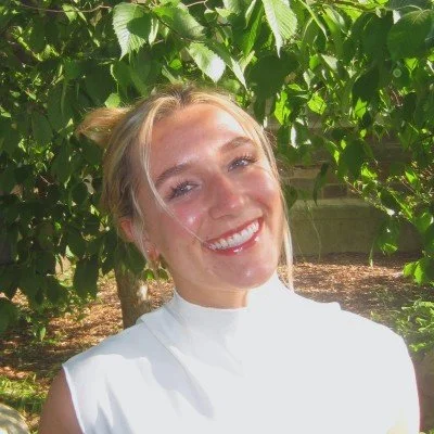 A woman smiling outdoors with lush green foliage in the background.