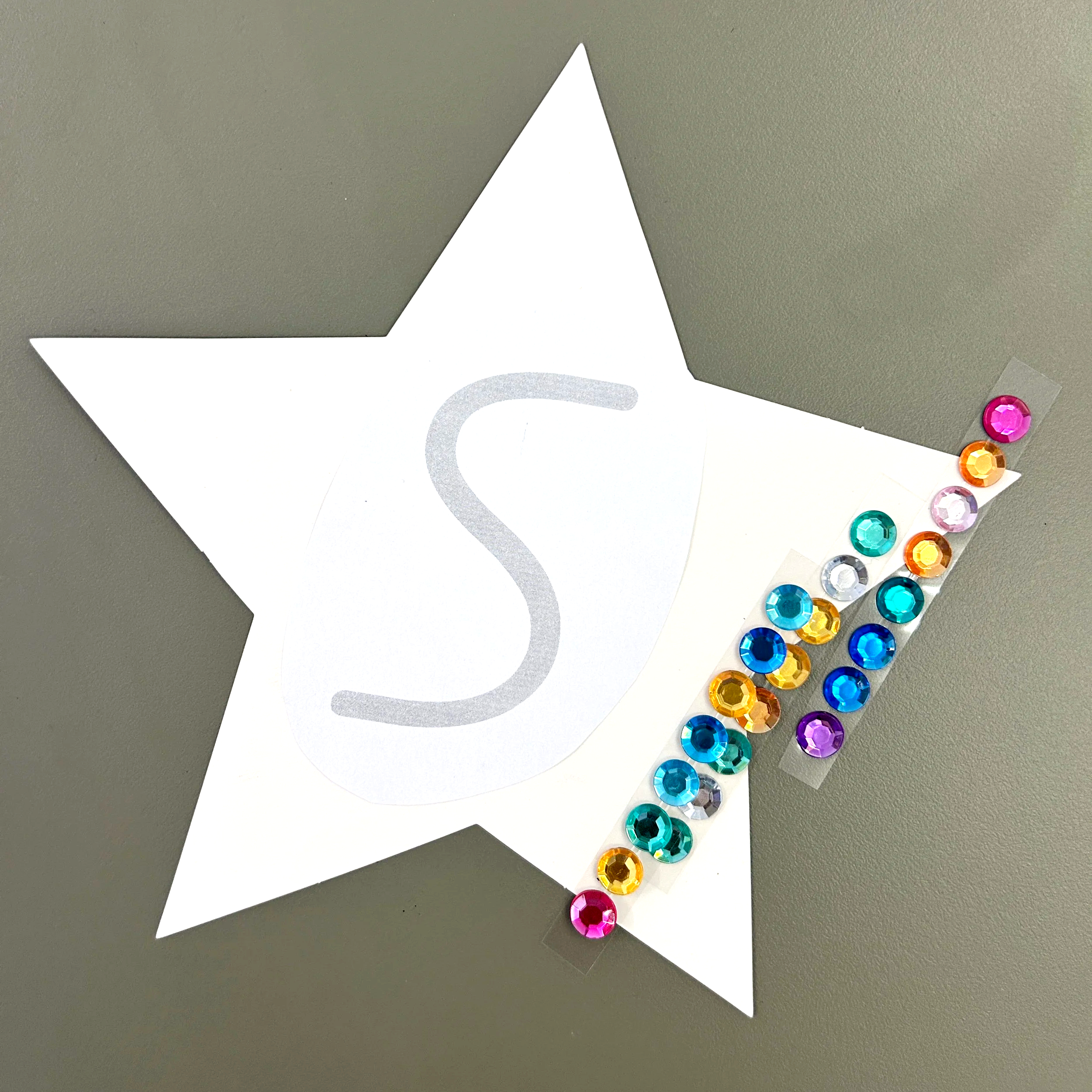 A group of young children sitting on a colorful alphabet rug, laughing and clapping while a female teacher interacts with them in a bright classroom decorated with educational posters and artwork.