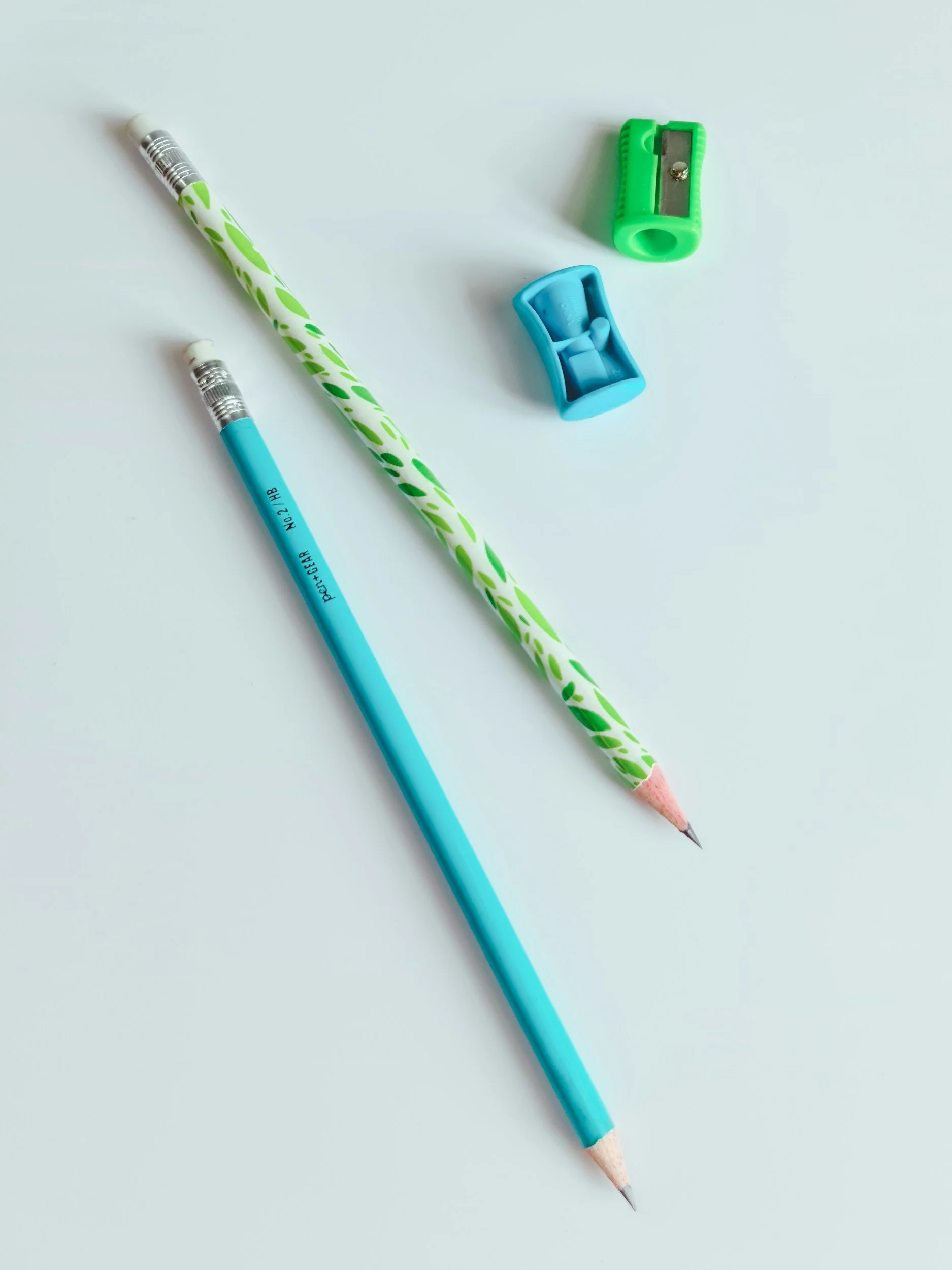 A light blue mechanical pencil with a pink tip, a green and white patterned pencil with a pink tip, a blue pencil sharpener, and a green utility knife on a white surface.