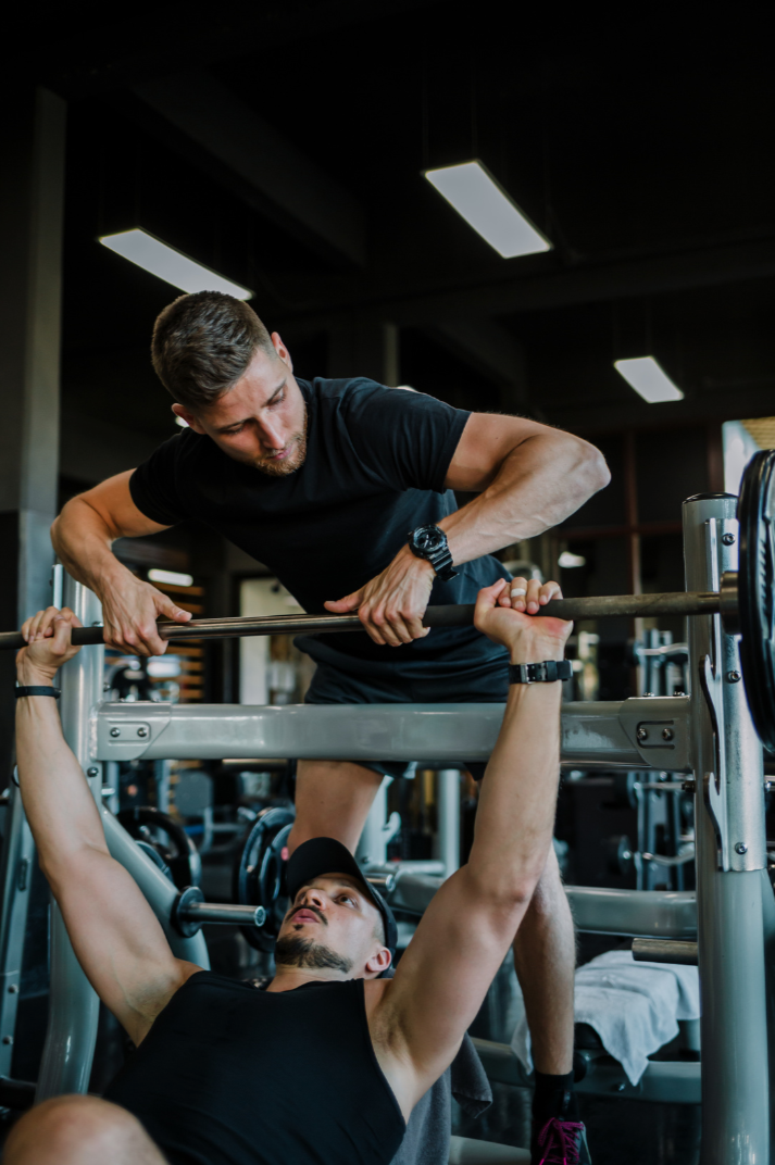 Two men working out in a gym; one is lying on a bench pressing a barbell, while the other assists by spotting him.
