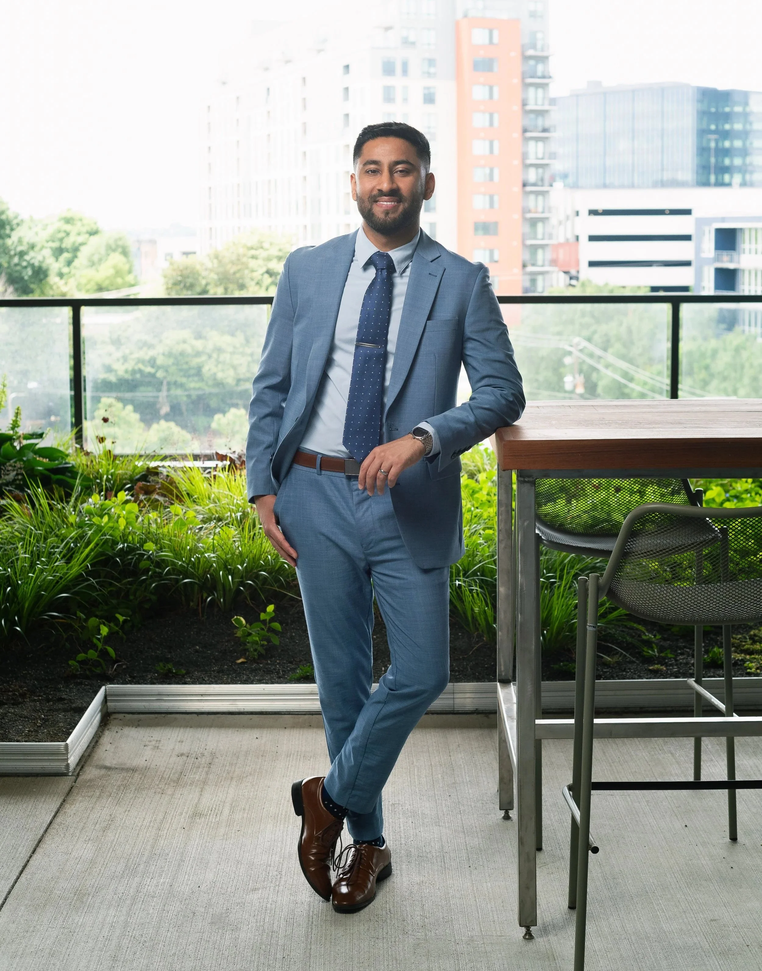 Anoop Takher in a light blue suit with a tie standing on a balcony with city buildings in the background.