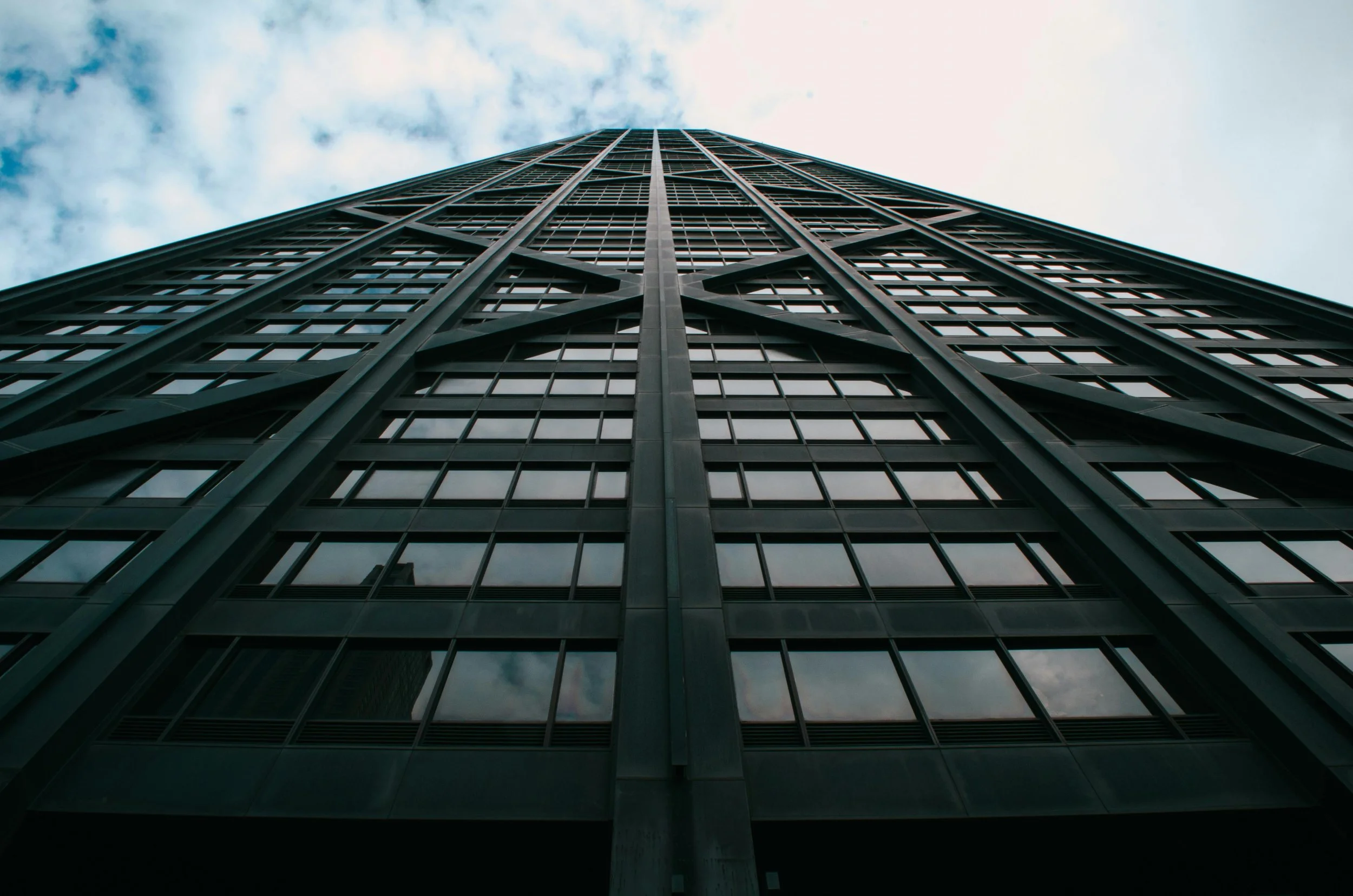 View of a tall modern skyscraper from the ground, emphasizing its glass windows and steel framework with a cloudy sky in the background.