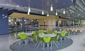 Modern cafeteria with green tables and chairs, interior lighting, and large windows.