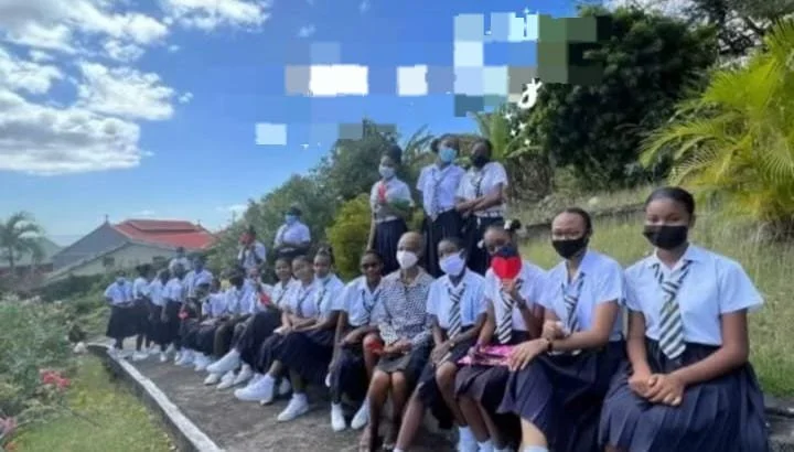 A group of students and teachers, all wearing face masks, sitting and standing on a grassy hill with plants and trees in the background, outside on a sunny day.