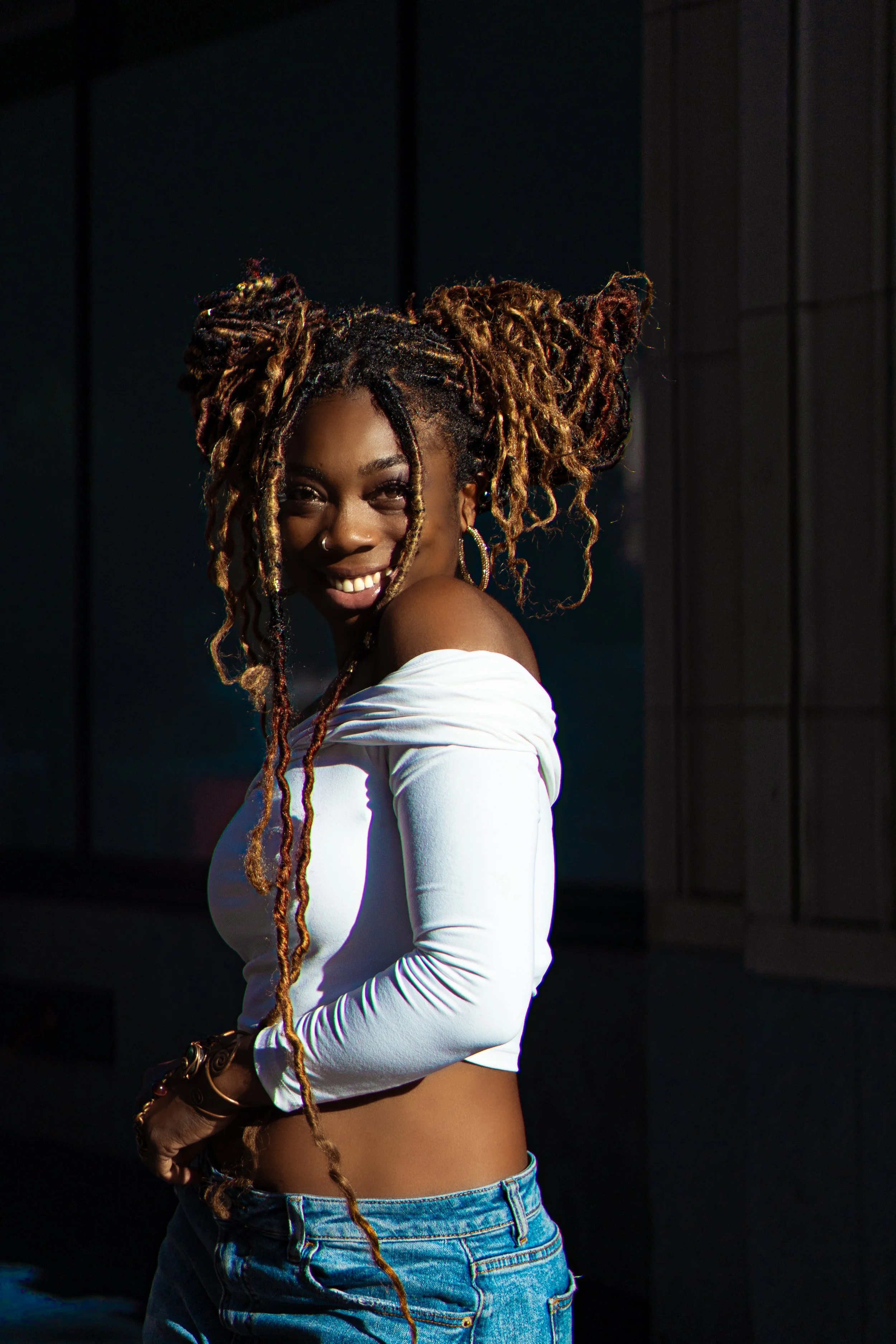 A smiling woman with dreadlocked hair wearing a white off-shoulder top and blue jeans, standing outdoors at sunset.