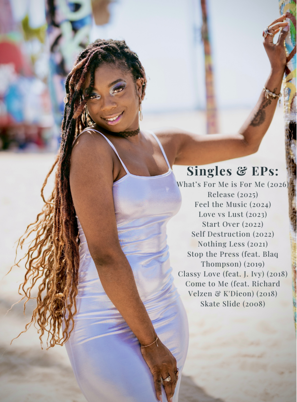 A woman with long, curly dreadlocks and a nose piercing wearing a white satin dress, standing outdoors on a beach with colorful umbrellas in the background. She is smiling and holding onto a painted wall, with a list of music singles and EPs by her s