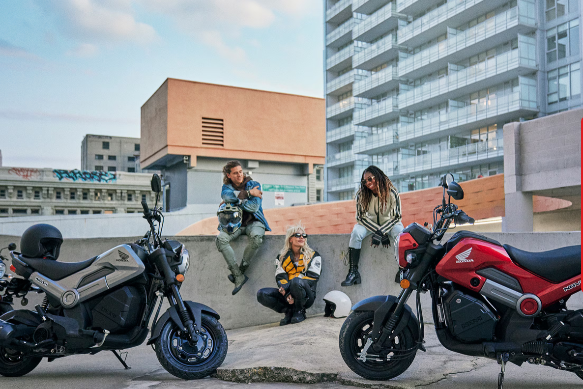 Four diverse women with motorcycles near urban buildings, some sitting on a ledge, one crouching with motorcycle helmet, during daytime.