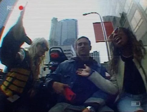 Three people sitting outdoors in an urban environment with tall buildings visible behind them.